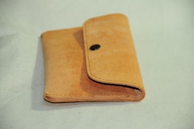 A light brown, leather wallet with a simple design and a metal snap closure. The wallet is positioned on a light surface and appears slightly worn, displaying a natural texture.