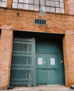 A brick industrial building facade with a steel folding gate and large green doors featuring signs that read 'Do Not Enter, Loading Unloading Only'. A traditional lamp is mounted above the entrance, and large windows are visible at the top.