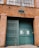 A brick industrial building facade with a steel folding gate and large green doors featuring signs that read 'Do Not Enter, Loading Unloading Only'. A traditional lamp is mounted above the entrance, and large windows are visible at the top.