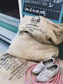 A collection of burlap sacks labeled with organic cocoa beans, a pair of rustic wooden clogs, and a coil of pink rope are placed near a storefront. A chalkboard on the wall advertises cold chocolate milk and ice cream.