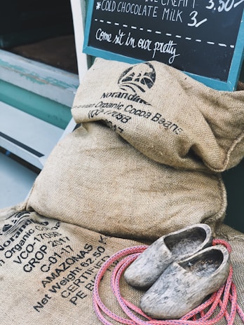 A collection of burlap sacks labeled with organic cocoa beans, a pair of rustic wooden clogs, and a coil of pink rope are placed near a storefront. A chalkboard on the wall advertises cold chocolate milk and ice cream.