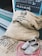 A collection of burlap sacks labeled with organic cocoa beans, a pair of rustic wooden clogs, and a coil of pink rope are placed near a storefront. A chalkboard on the wall advertises cold chocolate milk and ice cream.