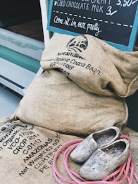 A collection of burlap sacks labeled with organic cocoa beans, a pair of rustic wooden clogs, and a coil of pink rope are placed near a storefront. A chalkboard on the wall advertises cold chocolate milk and ice cream.