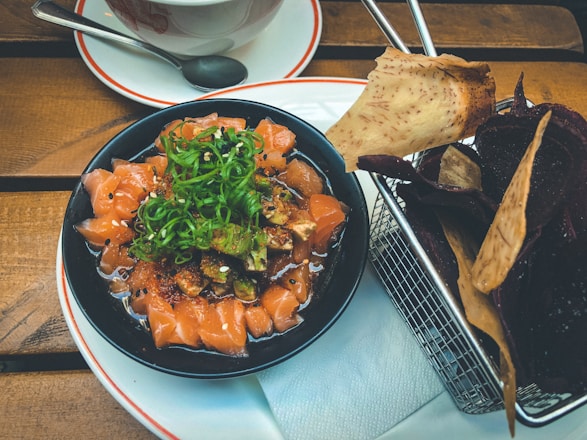 A bowl of fresh, finely diced salmon sashimi topped with sliced green onions and sesame seeds is served on a wooden table. Next to it is a metal basket containing assorted vegetable chips, including slices of taro and beetroot. A cup with a saucer and spoon is partially visible in the background.