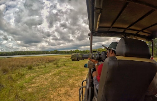 A person is seated in an open vehicle, using a large camera with a telephoto lens to photograph wildlife. The scene includes grassy plains under a dramatic, cloudy sky, with a dense tree line in the distance.