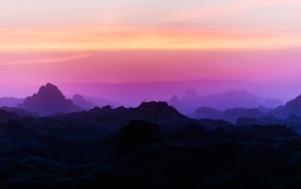 A serene landscape with rolling hills silhouetted against a vibrant sky. The horizon features gradients of orange to pink and purple, fading into soft blues as it meets the darkened terrain. The light creates a tranquil atmosphere.
