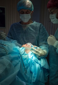A medical scene with a surgical team performing an operation. Two surgeons in scrubs, masks, and gloves are focused on the patient, who is lying on the operating table covered in blue surgical drapes. Medical instruments and bright lighting are visible, emphasizing the sterile environment.