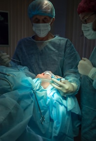 A medical scene with a surgical team performing an operation. Two surgeons in scrubs, masks, and gloves are focused on the patient, who is lying on the operating table covered in blue surgical drapes. Medical instruments and bright lighting are visible, emphasizing the sterile environment.