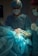 A medical scene with a surgical team performing an operation. Two surgeons in scrubs, masks, and gloves are focused on the patient, who is lying on the operating table covered in blue surgical drapes. Medical instruments and bright lighting are visible, emphasizing the sterile environment.