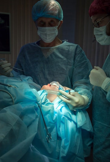 A medical scene with a surgical team performing an operation. Two surgeons in scrubs, masks, and gloves are focused on the patient, who is lying on the operating table covered in blue surgical drapes. Medical instruments and bright lighting are visible, emphasizing the sterile environment.