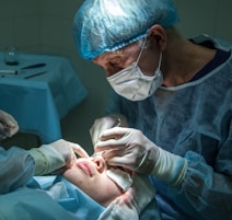 A medical professional is performing a surgical procedure on a patient's nose. Both individuals are wearing sterile attire, including gloves and masks. The surgeon focuses intently while using surgical instruments. The environment is clinical, with the patient lying down and partially covered.