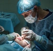 A medical professional is performing a surgical procedure on a patient's nose. Both individuals are wearing sterile attire, including gloves and masks. The surgeon focuses intently while using surgical instruments. The environment is clinical, with the patient lying down and partially covered.