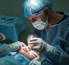 A medical professional is performing a surgical procedure on a patient's nose. Both individuals are wearing sterile attire, including gloves and masks. The surgeon focuses intently while using surgical instruments. The environment is clinical, with the patient lying down and partially covered.