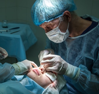 A medical professional is performing a surgical procedure on a patient's nose. Both individuals are wearing sterile attire, including gloves and masks. The surgeon focuses intently while using surgical instruments. The environment is clinical, with the patient lying down and partially covered.