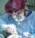 A healthcare professional wearing a red surgical cap, mask, and gloves is performing an intricate procedure. The person is using surgical tools and wearing specialized binoculars to enhance vision. The setting appears to be a brightly lit operating room.