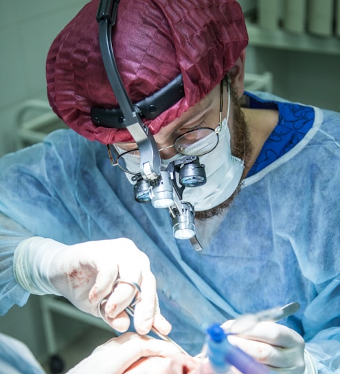 A healthcare professional wearing a red surgical cap, mask, and gloves is performing an intricate procedure. The person is using surgical tools and wearing specialized binoculars to enhance vision. The setting appears to be a brightly lit operating room.