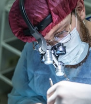 A focused medical professional wearing a red surgical cap and eyeglasses is intently working, equipped with surgical loupes for magnification. The person is in a blue sterile gown and wearing a white surgical mask, highlighting their attention and precision while performing a task.