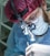 A focused medical professional wearing a red surgical cap and eyeglasses is intently working, equipped with surgical loupes for magnification. The person is in a blue sterile gown and wearing a white surgical mask, highlighting their attention and precision while performing a task.