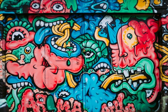 A vibrant and chaotic street art mural featuring a variety of exaggerated cartoonish faces and abstract shapes. The figures are intertwined, with wide eyes, open mouths, and bold expressions, blending into each other with surreal elements like tentacles, tongues, and dripping textures.