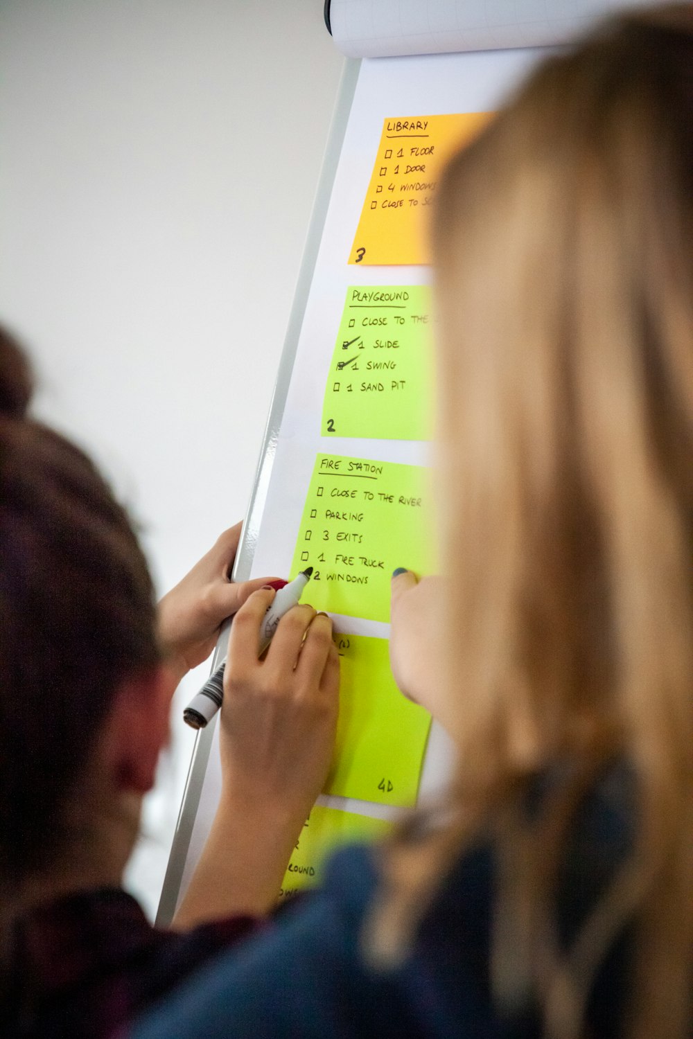 woman pointing yellow sticky paper with person holding pen and writing on paper on whiteboard