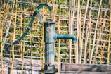 A vintage hand-operated water pump with a dark green color is set against a background of bamboo sticks and a wire fence. The pump's metallic surface shows signs of wear, suggesting it has been exposed to the elements. The overall setting has a rustic, countryside feel.