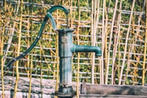 A vintage hand-operated water pump with a dark green color is set against a background of bamboo sticks and a wire fence. The pump's metallic surface shows signs of wear, suggesting it has been exposed to the elements. The overall setting has a rustic, countryside feel.