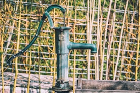 A vintage hand-operated water pump with a dark green color is set against a background of bamboo sticks and a wire fence. The pump's metallic surface shows signs of wear, suggesting it has been exposed to the elements. The overall setting has a rustic, countryside feel.