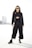 A woman is standing confidently in an open area with a cityscape in the background. She is wearing a stylish outfit, including a black blazer, cropped top, wide-leg pants, and ankle boots. Her long hair is loose, and she is accessorizing with dark sunglasses.