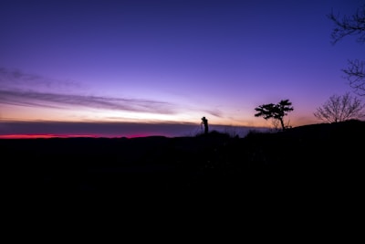 A striking silhouette of a person with a camera on a tripod captures a serene landscape during twilight. The sky transitions from a deep blue at the top to soft purple and pink hues near the horizon. Sparse trees are silhouetted against the colorful background, adding depth and contrast to the scene.