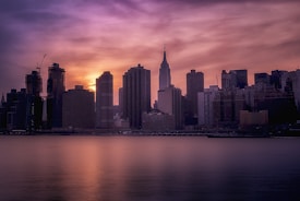 A city skyline silhouette with tall buildings against a colorful sunset sky. The water in the foreground reflects the warm hues of the setting sun, while the sky transitions from orange and pink to darker purple shades.