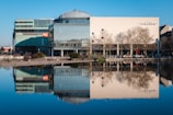 A large, modern building with glass facades and a dome-like roof is reflected in a calm body of water. Leafless trees line the area in front of the building, and cars are parked along the street. A few people are visible walking nearby.