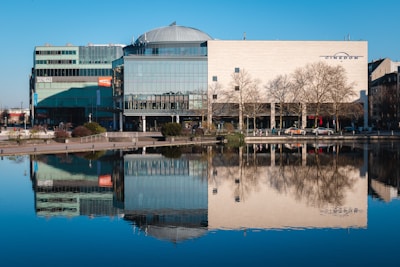 A large, modern building with glass facades and a dome-like roof is reflected in a calm body of water. Leafless trees line the area in front of the building, and cars are parked along the street. A few people are visible walking nearby.