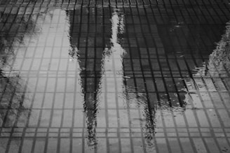 A black and white photograph capturing the reflection of a building with spires in a puddle on a tiled surface. The grid-like pattern of the tiles is prominent, with ripples in the water adding texture. The overall atmosphere is contemplative and artistic.