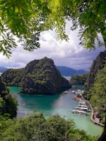 An idyllic tropical scene featuring a bay with turquoise waters surrounded by lush, green-covered limestone cliffs. Several boats are docked along a wooden pier leading out from the shore, and tree branches with green leaves frame the top part of the image.