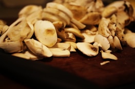 A pile of freshly sliced mushrooms sits on a dark wooden surface. The mushrooms are pale with varying shades of white and light brown, showcasing their fresh texture and natural patterns.