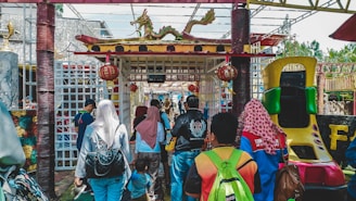 A group of people walking towards an entrance decorated with traditional Chinese architectural elements, including a dragon figure on top and red lanterns. The area appears festive and colorful, suggesting a possible cultural celebration or theme park setting. The crowd is diverse, wearing casual clothing and carrying bags. On the right, a slide designed for children is visible.
