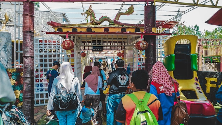A group of people walking towards an entrance decorated with traditional Chinese architectural elements, including a dragon figure on top and red lanterns. The area appears festive and colorful, suggesting a possible cultural celebration or theme park setting. The crowd is diverse, wearing casual clothing and carrying bags. On the right, a slide designed for children is visible.