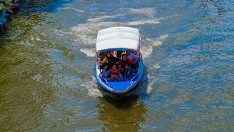 A small motorized boat navigates through a body of water with several people on board, including a child. The boat has a blue hull and a white canopy. The water is slightly choppy, creating light ripples and wakes behind the boat. The image captures a sense of movement and outdoor activity.
