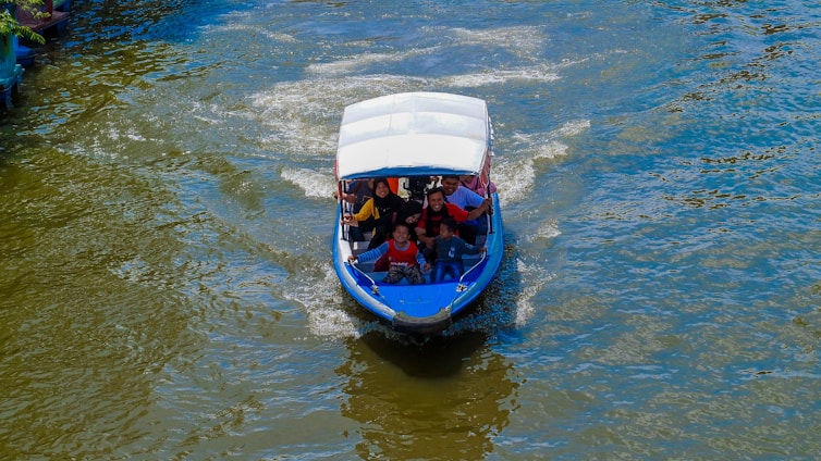 A small motorized boat navigates through a body of water with several people on board, including a child. The boat has a blue hull and a white canopy. The water is slightly choppy, creating light ripples and wakes behind the boat. The image captures a sense of movement and outdoor activity.