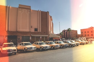 A row of classic beige and white taxis is parked along a street with an art deco-style building in the background. The scene takes place in an urban area with palm trees and several people gathered near the vehicles. The lighting suggests a warm, sunny day.