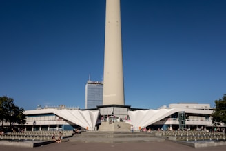 A tall concrete tower rises prominently into the sky with a clear blue backdrop. At the base, a modern, angular building with white, geometric structures serves as its foundation. People are scattered around the open plaza in front of the building, enjoying the sunny day. A nearby skyscraper features the name 'Park Inn' at its top. Small trees frame the scene, adding greenery to the urban landscape.