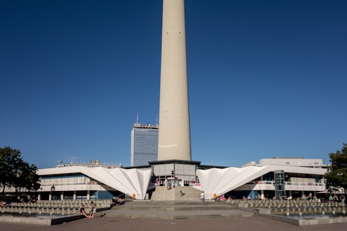 A tall concrete tower rises prominently into the sky with a clear blue backdrop. At the base, a modern, angular building with white, geometric structures serves as its foundation. People are scattered around the open plaza in front of the building, enjoying the sunny day. A nearby skyscraper features the name 'Park Inn' at its top. Small trees frame the scene, adding greenery to the urban landscape.