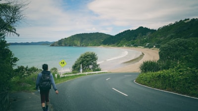 A lone person with a backpack is walking along a curved road by the beach. The sea is calm with turquoise and blue colors blending together, while the beach is sandy and stretches towards distant green hills. There are a lot of green trees and vegetation on either side of the road. A yellow caution sign is seen near the side of the road. The sky is partly cloudy.