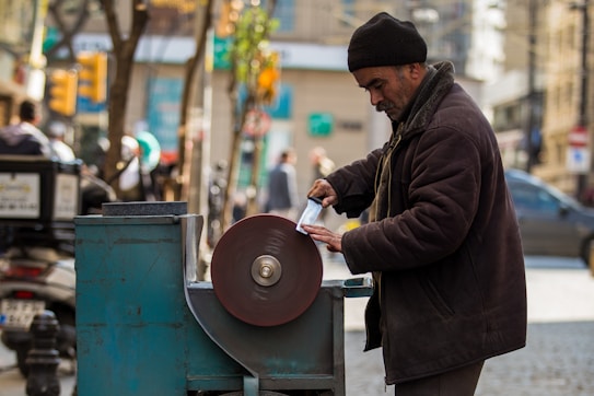 A man in warm clothing is sharpening a knife on a street-side grinding wheel in an urban setting. The backdrop includes blurred figures of passersby, vehicles, and buildings, with a soft focus that highlights the main subject.
