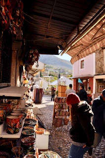 A narrow street with cobblestone pavement features various market stalls displaying colorful ceramics and textiles. People are browsing the merchandise, and a mountainous landscape can be seen in the background.