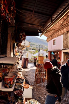 A narrow street with cobblestone pavement features various market stalls displaying colorful ceramics and textiles. People are browsing the merchandise, and a mountainous landscape can be seen in the background.