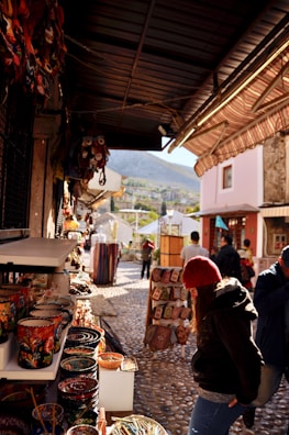 A narrow street with cobblestone pavement features various market stalls displaying colorful ceramics and textiles. People are browsing the merchandise, and a mountainous landscape can be seen in the background.
