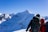 Smiling couple standing on a mountain trail with backpacks, surrounded by snow-capped peaks under a clear blue sky.