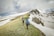 Hikers trek along a snow-covered mountain ridge, surrounded by patches of green grass dotted with yellow flowers. The sky is overcast with thick clouds, casting a muted light over the scene. The hikers wear backpacks and are bundled for cold weather, suggesting a challenging outdoor adventure.