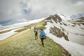 Hikers trek along a snow-covered mountain ridge, surrounded by patches of green grass dotted with yellow flowers. The sky is overcast with thick clouds, casting a muted light over the scene. The hikers wear backpacks and are bundled for cold weather, suggesting a challenging outdoor adventure.